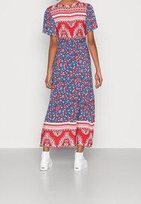 Maxiklänning med korta ärmar, med en blå bas, röda blommönster och en dekorativ röd och vit fåll. Kombineras med vita platåskor.