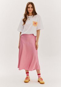 Biela krátkym rukávom tričko s dizajnom vrecka broskyne a jahody, skombinované s ružovou saténovou maxi sukňou a béžovými teniskami.