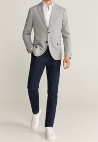 Blazer grigio su misura con rever a lancia, abbinato a jeans scuri e sneakers bianche. Il blazer presenta tasche frontali e un solo bottone.