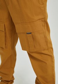 Bruna cargobyxor i texturerat tyg, med stora sidofickor, rak passform och en liten märkesskylt nära midjan.