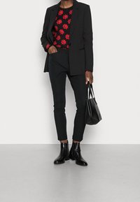 Schwarze Blazer über einem rot blumigen Oberteil, kombiniert mit schwarzen Hosen und Stiefeletten. Accessoires mit einer schwarzen Handtasche.