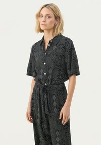 Black short-sleeve button-up shirt with a collar, featuring white geometric and floral patterns, paired with a tied waist, flowing design.