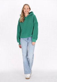 Grøn fleece hoodie med en struktureret overflade, oversized ærmer og en kængurulomme, båret med lyseblå flared jeans og hvide sneakers.