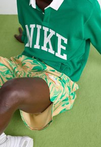 Grön sweatshirt med vit "NIKE" text, i kombination med gula shorts med gröna marmor mönster. Slät textur, avslappnad design.