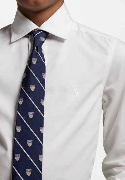 Camisa blanca con cuello puntiagudo, que presenta un pequeño logo bordado. Corbata azul marino con rayas diagonales y acentos con patrón de escudo.