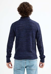 Suéter de punto azul marino con cuello alto y mangas largas, que presenta puños acanalados y patrones texturizados, combinado con jeans de color azul claro.