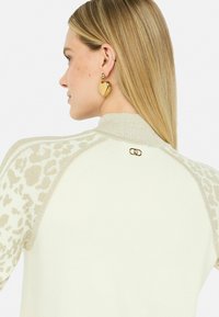 Maglione color crema con dettagli metallici testurizzati e maniche a stampa leopardata. Presenta un colletto a coste e un logo dorato sul retro.