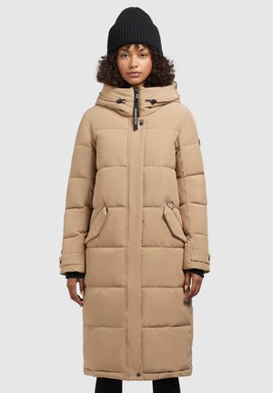 Doudoune beige avec capuche, motif matelassé, fermeture éclair, deux poches latérales et accents noirs ; portée avec un bonnet en tricot noir.