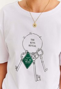 Biała koszulka z nadrukiem breloka, na którym wiszą cztery vintage klucze oraz zielona metka z napisem "Miasto nad morzem od 1980".
