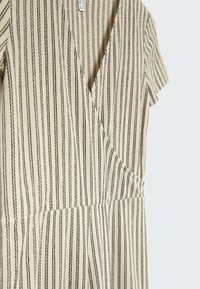 Robe cache-cœur beige avec des rayures noires verticales, manches courtes et taille cintrée. Tissu doux avec une texture lisse.