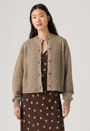 Young woman with long dark hair wears a textured beige cardigan over a brown dress with small light blue floral prints.