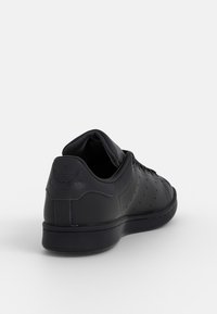 Černá kožená teniska s perforovaným zdobením, zaoblenou špičkou a logem na patě. Má plochou podrážku a polstrovaný lem pro pohodlí.