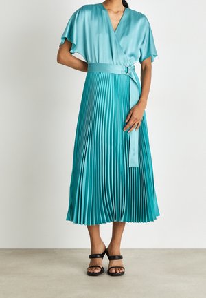 Vrouw die een teal satijnen wikkeljurk draagt met zwierige mouwen, geplooide midirok, bijpassende riem en zwarte hakken, staande op een grijze vloer.