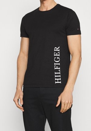 Muž nosící černé tričko s krátkým rukávem s bílým nápisem "HILFIGER" vertikálně na pravé straně a malým logem na levém rukávu.