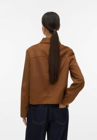 Veste en daim marron avec un col, des coutures aux épaules et une coupe courte. Associée à un jean bleu foncé. Texture lisse, détails minimalistes.