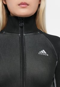 Mustan värinen urheilullinen vetoketjugaaleri, jossa on korkea kaulus, ribbed-pinta, valkoinen adidas-logo vasemmassa rinnassa ja kontrastiväriset ompelut.