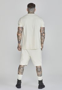 T-shirt beige a maniche corte e pantaloni corti abbinati, realizzati in tessuto morbido. Presenta una vestibilità comoda e tatuaggi visibili. Indossate scarpe nere alte.