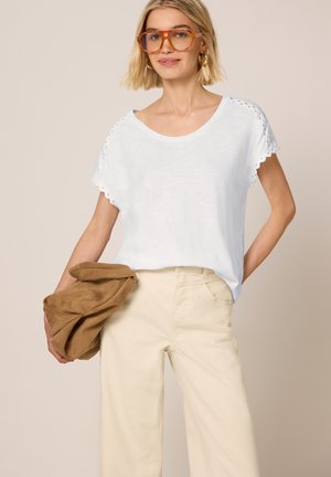 Femme aux cheveux blonds courts portant des lunettes à monture orange, un haut blanc à manches courtes, un pantalon crème, tenant un sac marron sur un fond neutre.