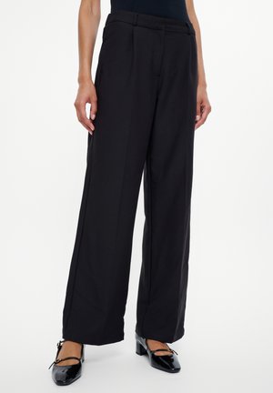 Zwarte wijde broek met een getailleerde pasvorm, voorzien van een gladde textuur en voorplooien. Gecombineerd met gepatenteerde leren schoenen met blokhak.