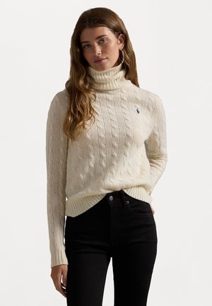 Maglione a collo alto color crema con motivo a trecce e orlo a costine. Presenta un piccolo logo blu sul petto. Abbinato a jeans neri.