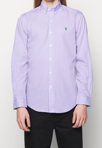 Man som bär en lila och vit vertikalt randig knapp-up skjorta med en grön broderad logotyp på vänster bröst, tillsammans med svarta byxor.