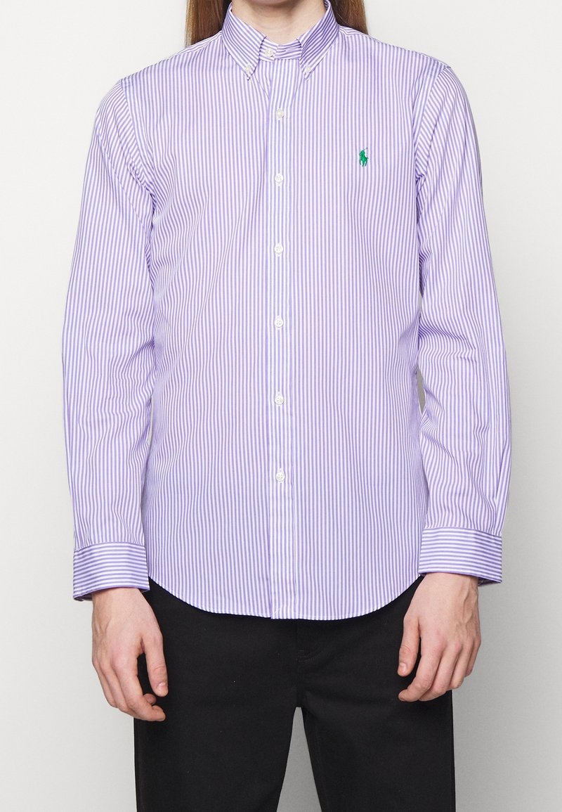 Man som bär en lila och vit vertikalt randig knapp-up skjorta med en grön broderad logotyp på vänster bröst, tillsammans med svarta byxor.