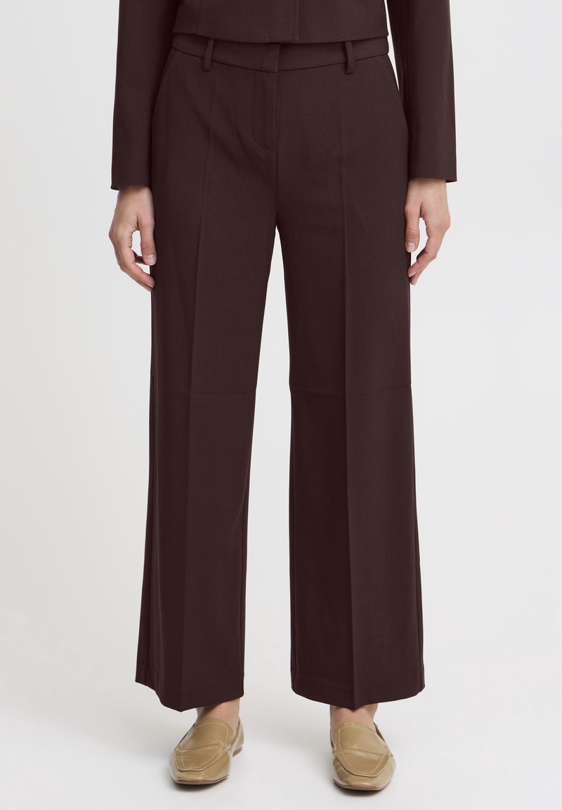 Femme portant un pantalon large marron foncé et des mocassins beiges, debout devant un fond clair uni.