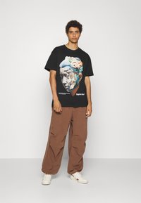 Sort kortærmet T-shirt med et grafisk billede af en klassisk skulptur i farverige nuancer. Båret med løse brune bukser og hvide sneakers.