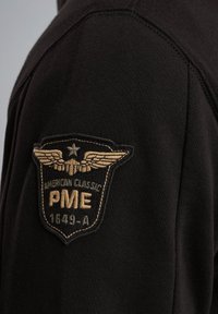 Zwarte sweatshirt met een gestructureerde stof, voorzien van een groot geborduurd patch in goud en zwart op de schouder, met vleugels en tekst.