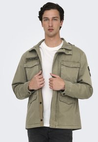 Giacca in cotone verde oliva con zip frontale, chiusure a bottone e multiple tasche. Presenta ricamo "U.S. ARMY" sul lato sinistro del petto.