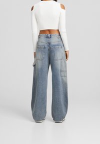 Pantalones de mezclilla de tiro alto y pierna ancha en azul claro con una textura descolorida. Cuenta con bolsillos tipo cargo y un ajuste suelto, combinados con una blusa corta de ribete blanca.