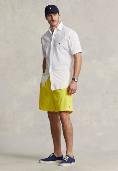 White short-sleeve button-up shirt, bright yellow shorts, navy cap, navy shoes, with a wristwatch, standing on a neutral background.