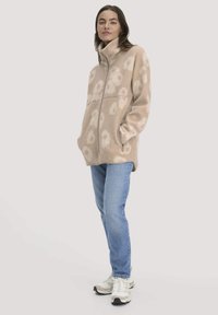 Vrouw in een beige gebloemde fleecejack, blauwe jeans en witte sneakers, met handen in de zakken staand tegen een effen achtergrond.