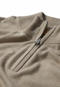 Suéter de pullover beige con cremallera, hecho de un suave tejido de punto, que cuenta con un cuello alto y una cremallera plateada, con una etiqueta de tela cerca de la abertura.