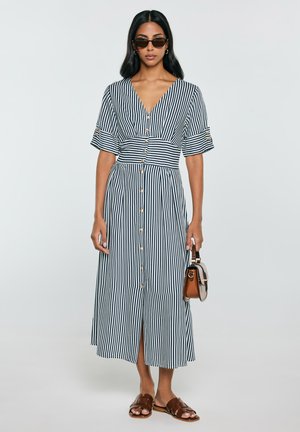 Kobieta ubrana w czarno-białą, pionowo paskowaną sukienkę midi z guzikami, brązowe sandały, okulary przeciwsłoneczne i trzyma małą torebkę.