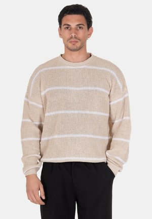 Uomo con capelli corti e scuri che indossa un maglione beige lavorato a maglia con righe orizzontali bianche e pantaloni neri, in piedi su sfondo bianco.