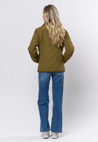 Olivzaļš kapucis ar dimanta rakstu, ar augsta apkakli. Valkāts kopā ar zilām džinsām un pelēkām sporta kurpēm, redzams no aizmugures.