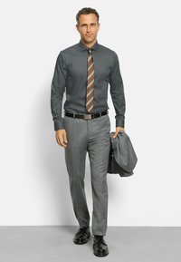 Herrenoutfit mit einem dunkel gemusterten Hemd, einer braunen gestreiften Krawatte, grauen gemusterten Hosen und schwarzen Schuhen. Ein grauer Blazer hängt über dem Arm.