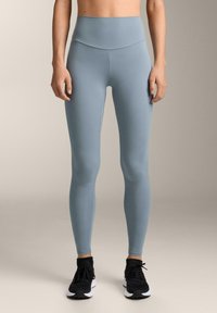 Vaaleansiniset korkeat legginsit, jotka on valmistettu sileästä ja venyvästä materiaalista, istuvat tiukasti sääressä, käytetään mustien urheilukenkien kanssa.