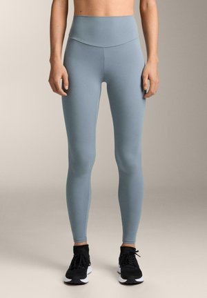 Világoskék, magas derekú leggings, amelyek sima, rugalmas anyagból készültek, testhez simuló szabásúak, fekete sportcipővel viselve.