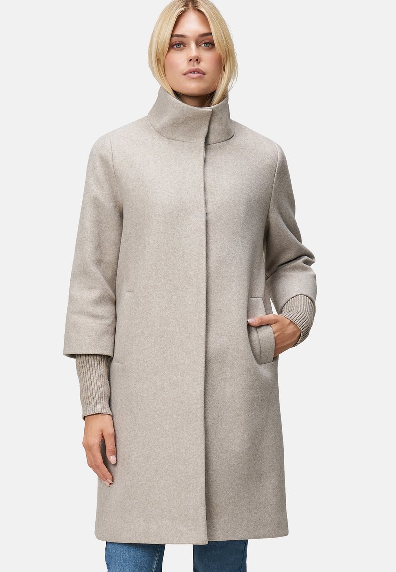 Manteau en laine beige clair avec un col haut, des manches larges et des poignets en tricot côtelé. Il dispose de poches latérales et d'une silhouette droite.