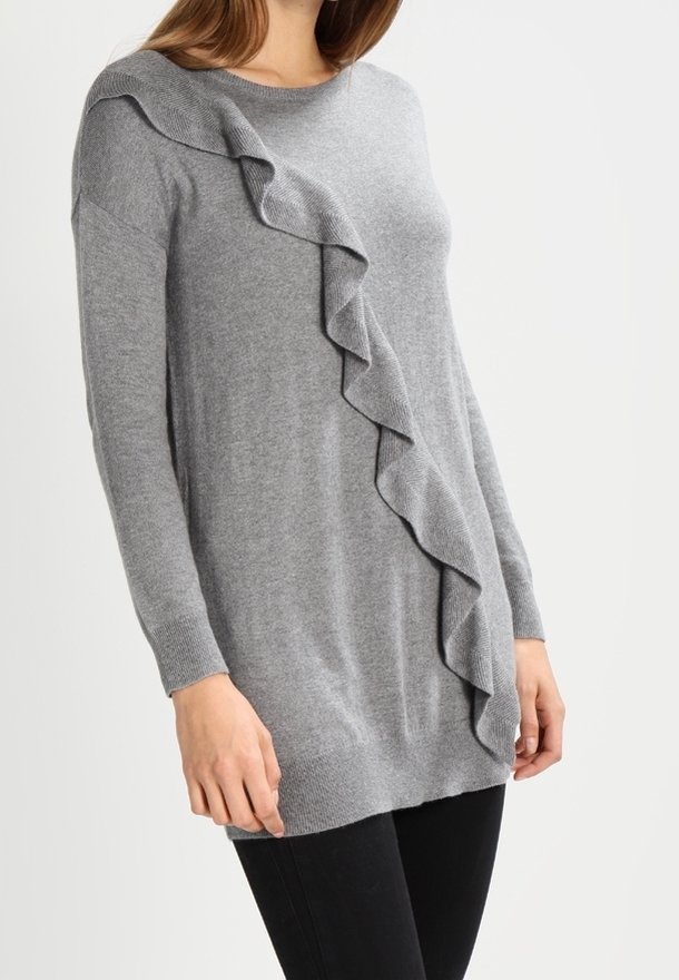Femme portant un pull en maille gris à manches longues avec un détail de volant diagonal de l'épaule gauche à l'ourlet droit, associé à un pantalon noir.