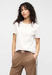 Λευκό βαμβακερό t-shirt με κοντά μανίκια, με ανάγλυφο σχέδιο λογότυπου στο κέντρο. Συνδυάζεται με καφέ παντελόνια και ορατό λογότυπο στη μέση.