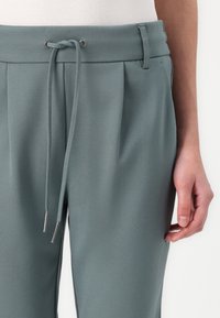 Hellgrüne Hose mit einem Kordelzugbund, Falten und metallischen Akzenten. Weicher Stoff, taillierte Passform und minimalistisches Design.