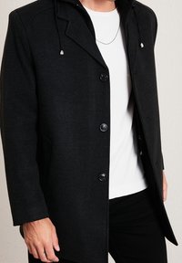 Cappotto in misto lana nero con design doppiopetto, bottoni prominenti e cappuccio, caratterizzato da una superficie liscia e tasche laterali nascoste.