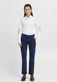 Weißes Hemd mit Knöpfen und floralen Stickereien an den Ärmeln, kombiniert mit marineblauen Hosen. Das Model steht in dunklen Schuhen vor einem schlichten Hintergrund.