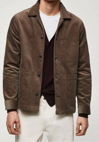 Uomo che indossa una giacca in velluto a coste marrone su un cardigan scuro abbottonato e pantaloni bianchi, su uno sfondo neutro.