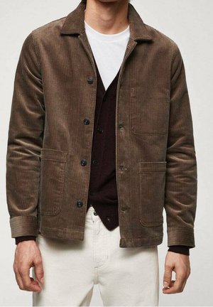 Man wearing a brown corduroy jacket over a dark buttoned cardigan and white pants against a plain background.