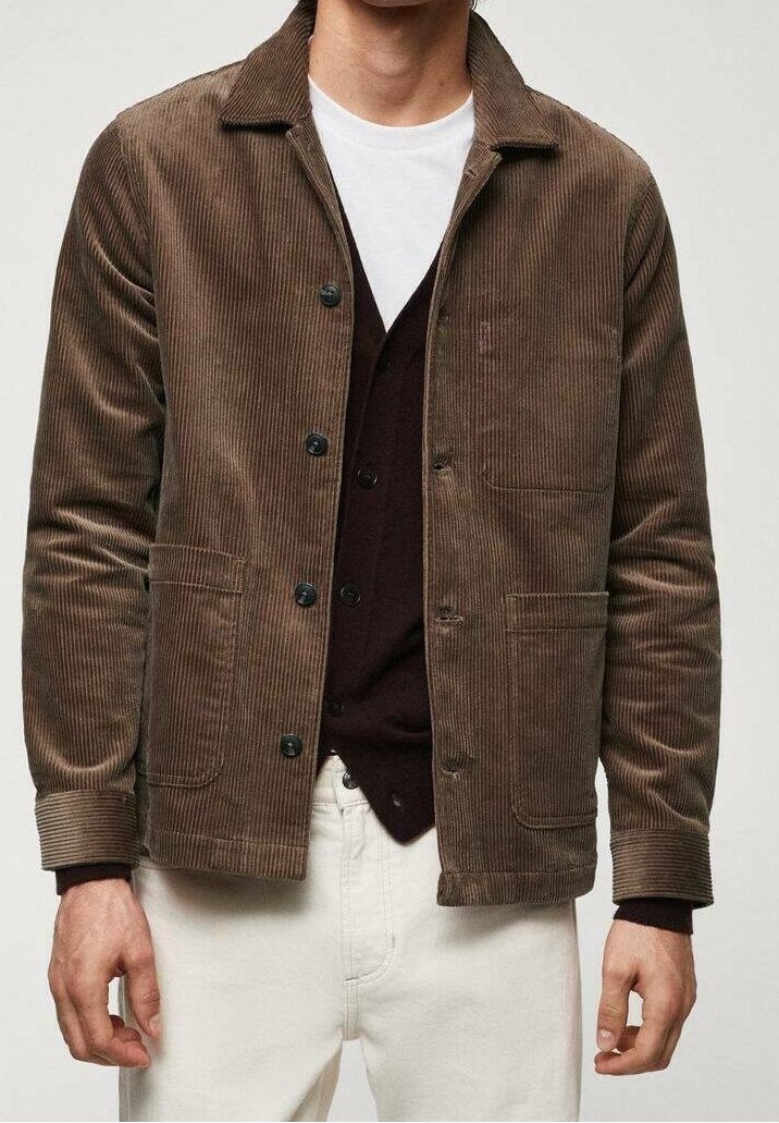 Uomo che indossa una giacca in velluto a coste marrone su un cardigan scuro abbottonato e pantaloni bianchi, su uno sfondo neutro.