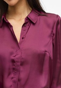 Camisa de seda ciruela con cuello tipo solapa, cierre de botones en la parte delantera y textura suave. Presenta costuras sutiles y un bolsillo en el pecho.